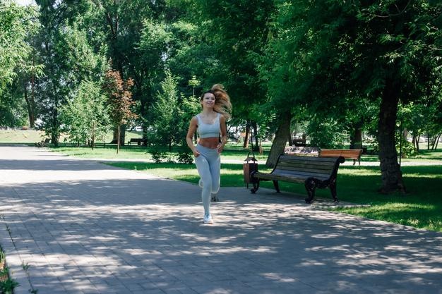 Running fast smiling young sporty woman warming up stretching in grey sportswear in park in sunny su...